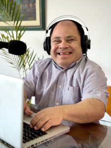 Shanir smiling in his office, using a laptop a micrphone and headphones-